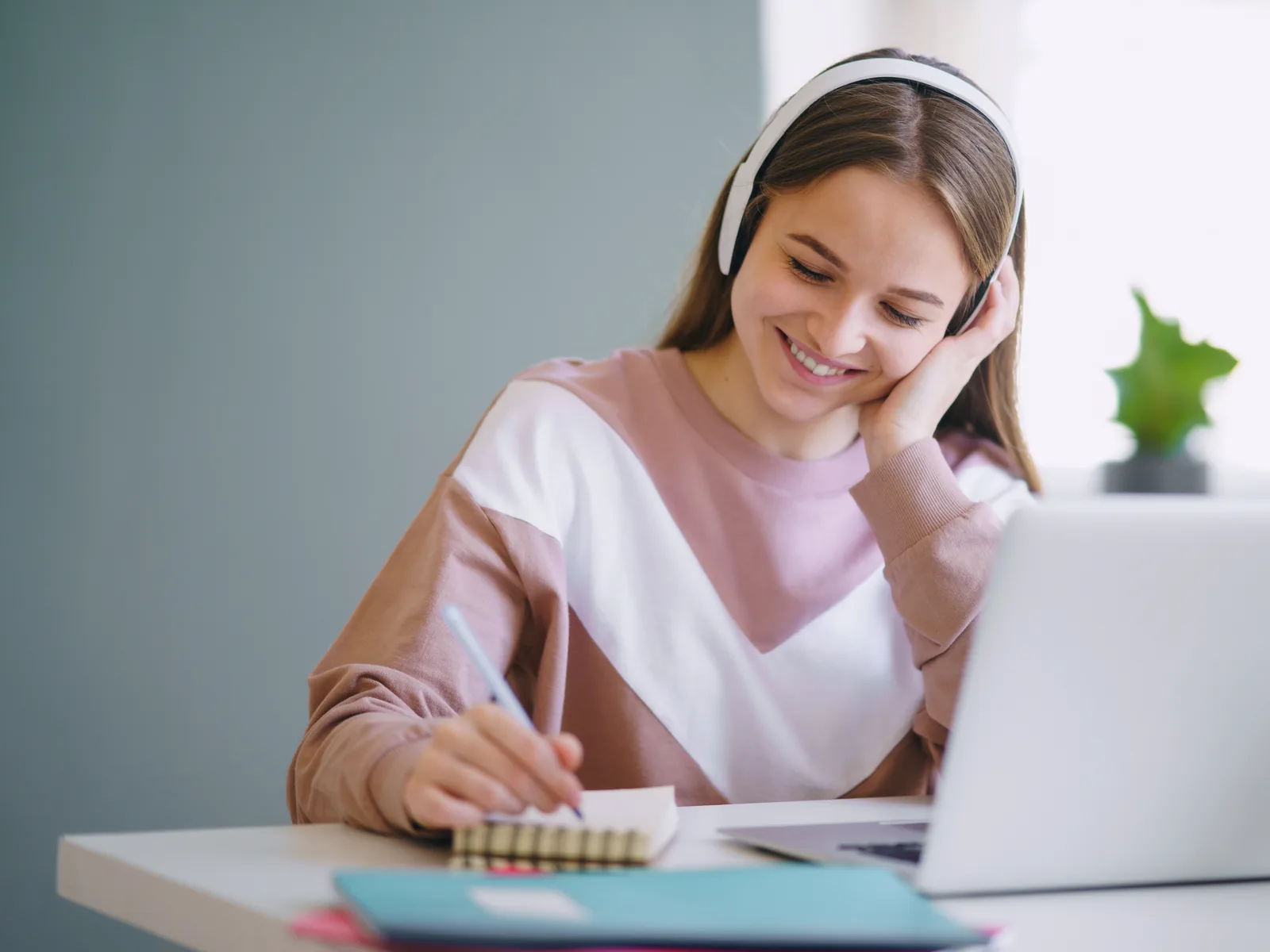 A student wearing headphones taking notes beside a laptop.